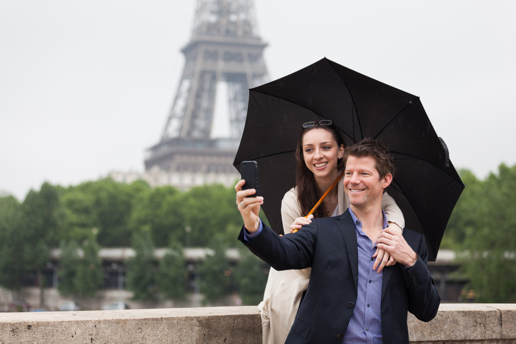 photo in bad weather of a smiling young couple under an umbrella in Paris with Eiffel Tower in the background