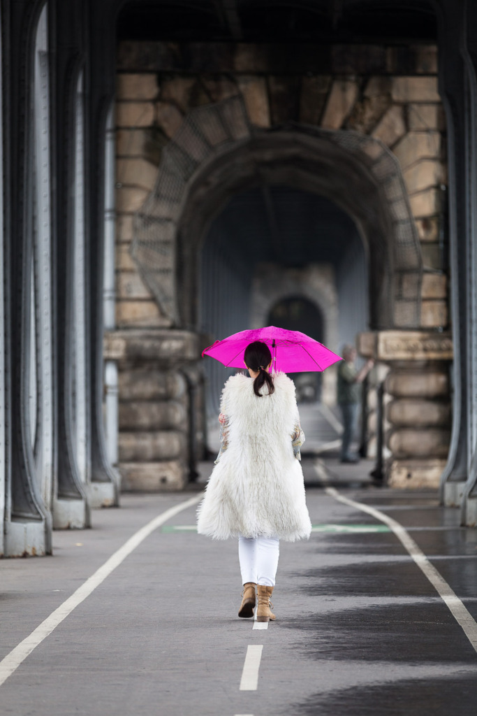 Photo in bad weather of a woman carrying a pink umbrella and wearing a fur coat walks away from the camera
