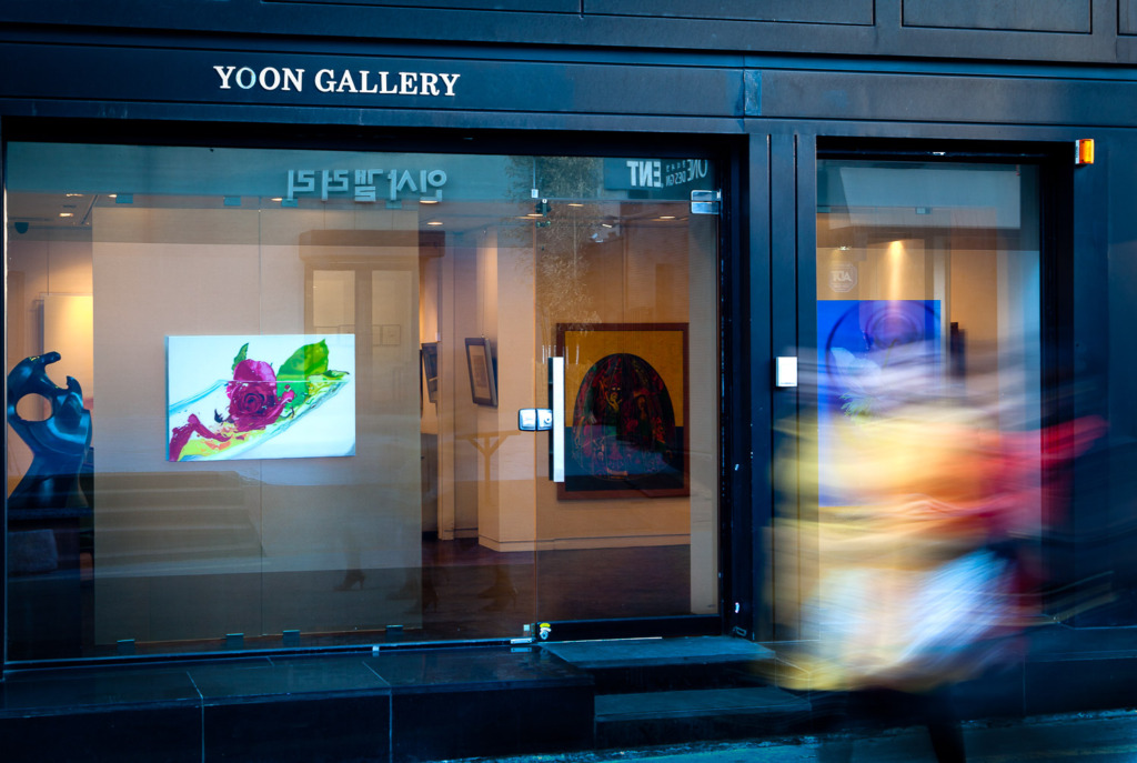 Add movement to your travel photo by shooting a long exposure photo of the window of an art galley in Seoul, South Korea with 3 women walking by.