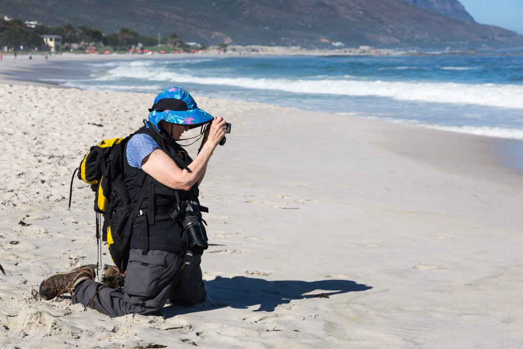 Older woman kneeling on the beach to take a photograph. She is wearing 2 cameras, a backpack, and a hat.