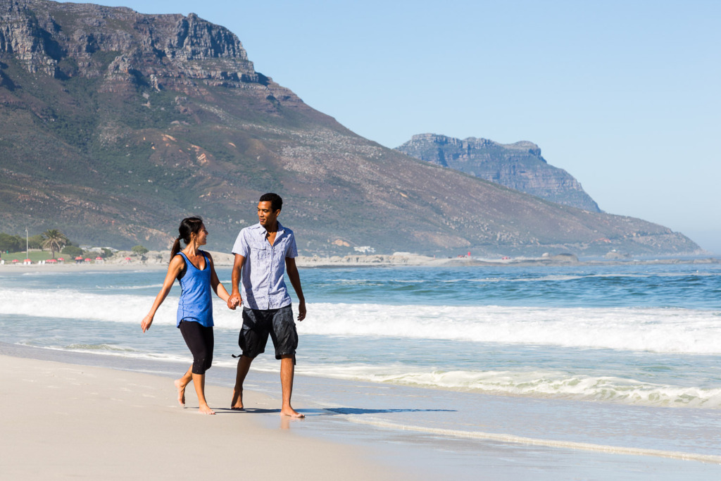 beach photo of couple walking in Camps Bay with mountains behind them.