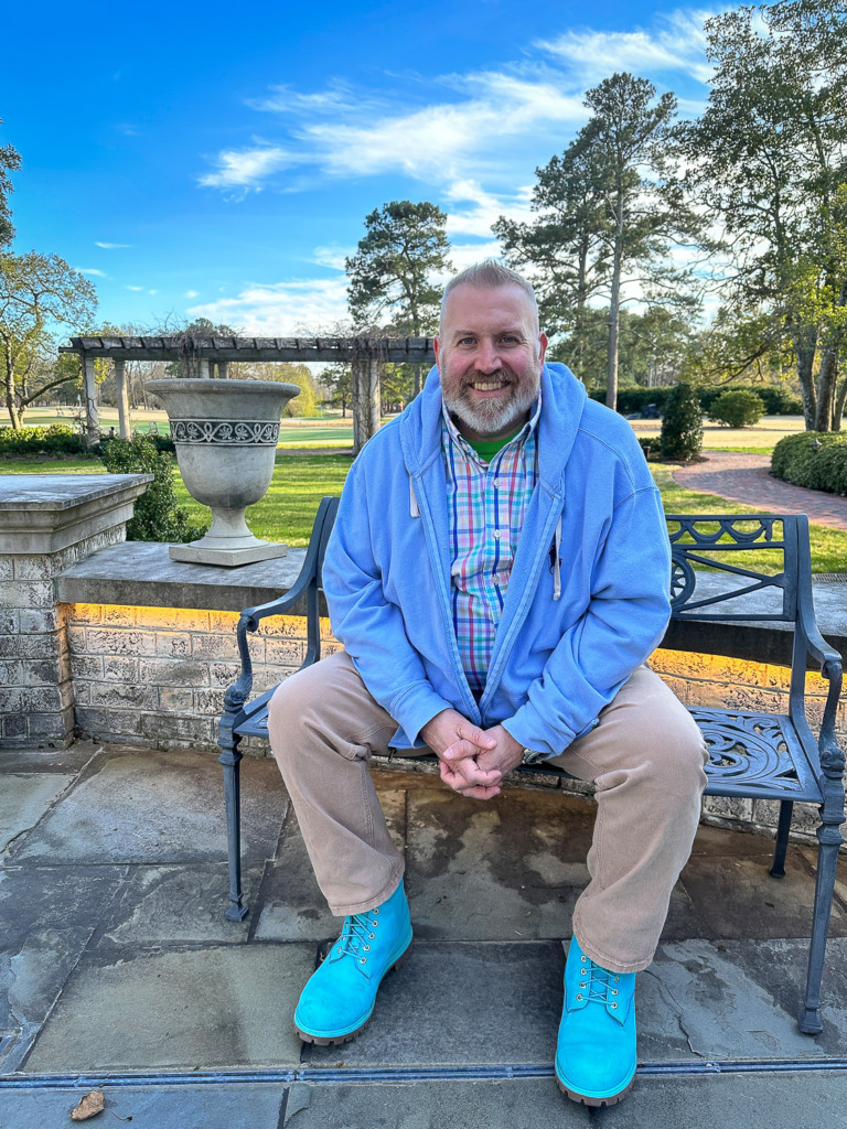 Pops of color can help with creative storytelling as in this portrait of smiling bearded man in light blue jacket, bright plaid shirt and turquoise blue shoes sitting on a garden bench with a garden behind him.