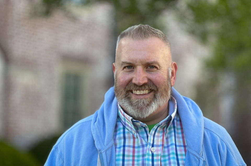 Portrait of smiling bearded man in light blue jacket and bright plaid shirt.