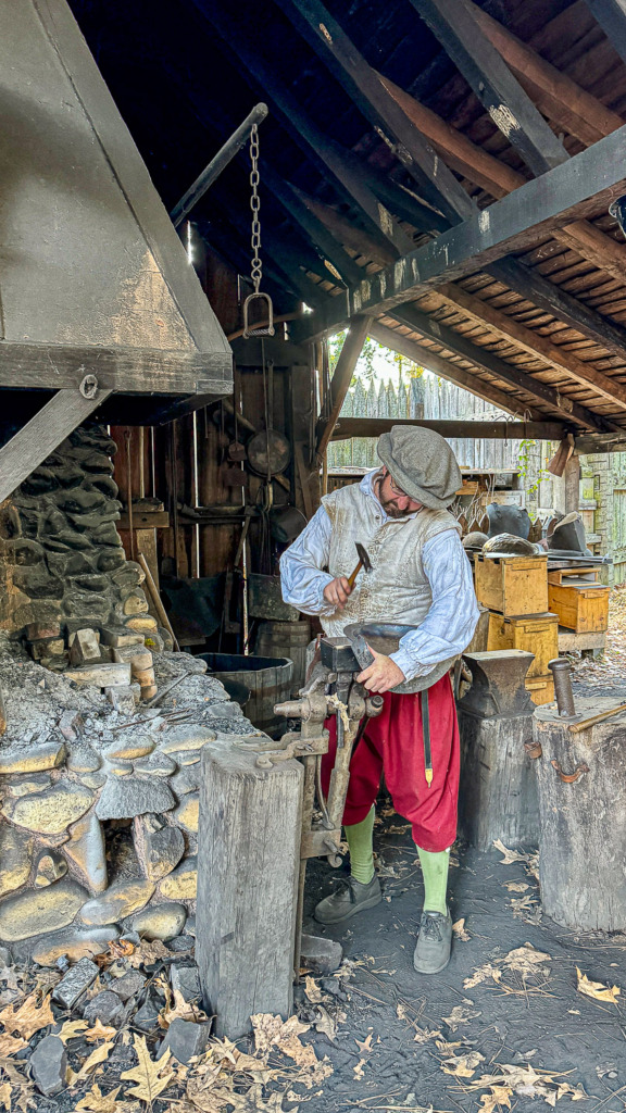 Photo of the blacksmith at James Fort in Jamestown Settlement, Virginia. He is wearing red pants, green socks and a white shirt and protective vest, plus a gray hat. He is using a hammer to shape a metal helmet.