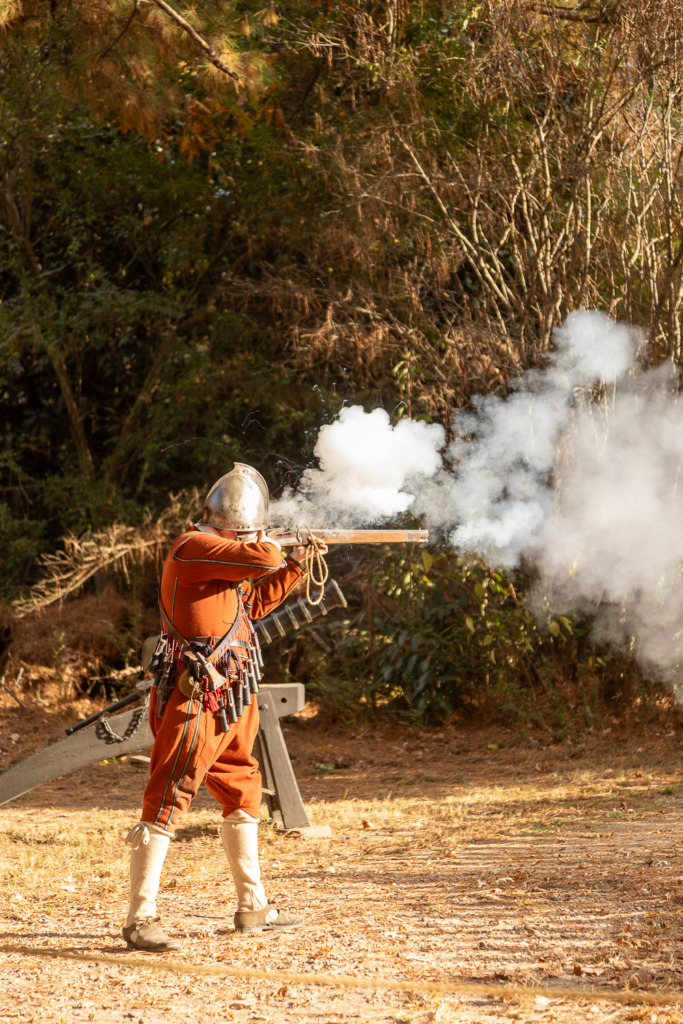 A historical interpreter firing a musket in Jamestown Settlement, Virginia. He is dressed in a red soldier's uniform and a metal helmet.