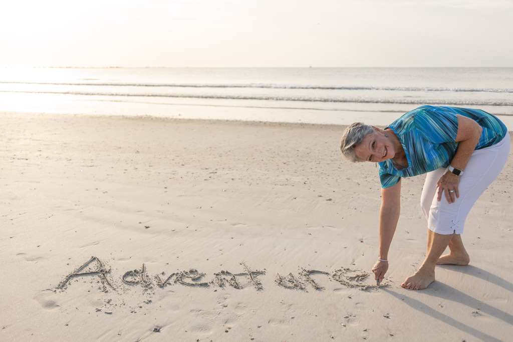 Photo of a smiling woman on a beach. She is writing Adventure in the sand. She's wearing white slacks and a striped blue shirt and is barefooted. Shooting a photo like this is like shooting a snow photo.