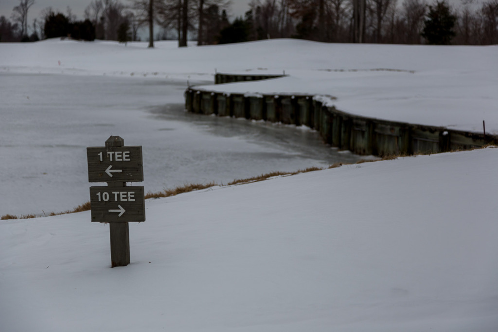 Snow photos can be very dark like this one of a sign at a golf course with snow on the ground and an icy pond behind it.